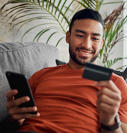 Man looking at his credit card while holding his phone on the couch at home.
